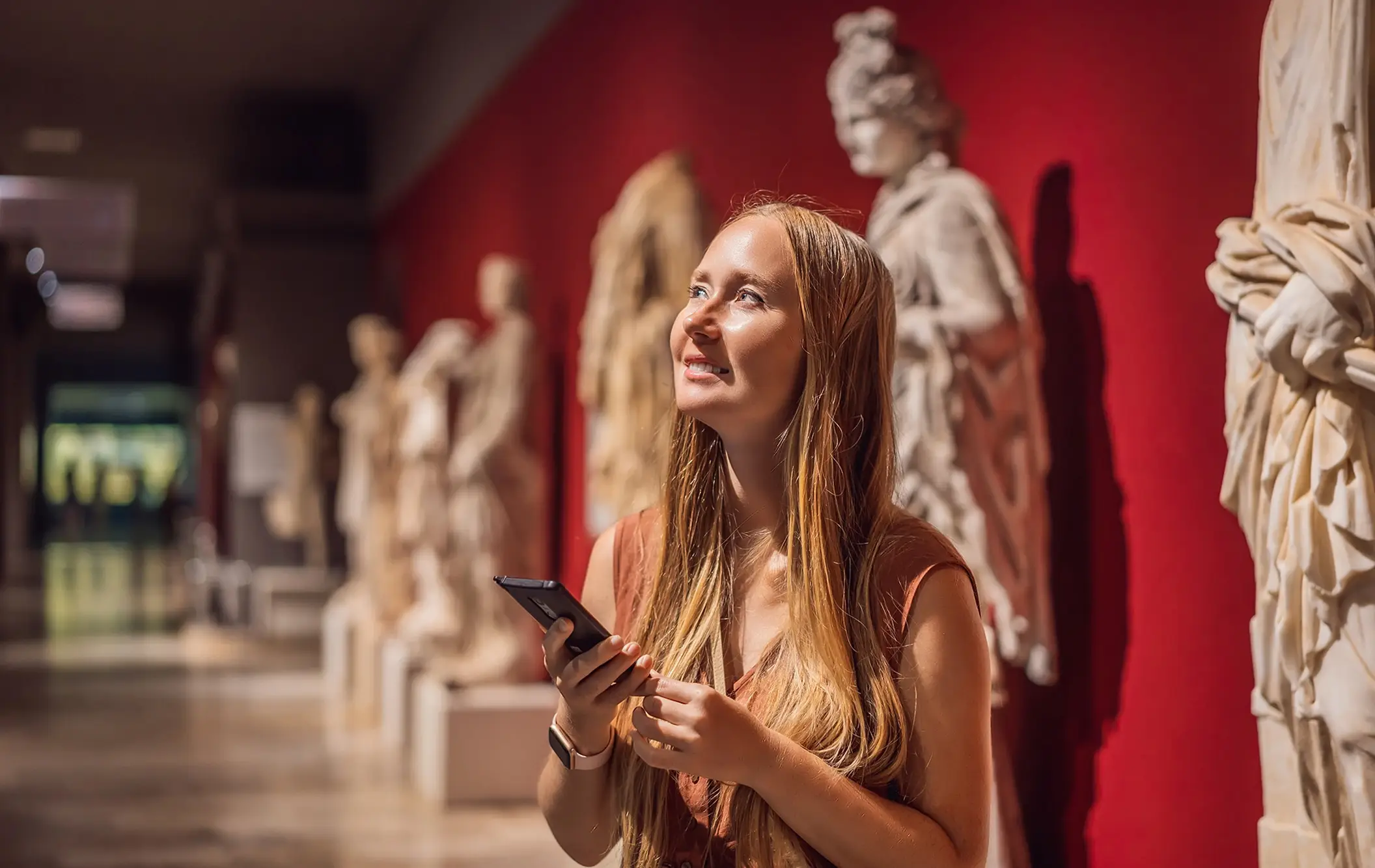 Young woman holding phone looking at sculptures in a museum with red walls.